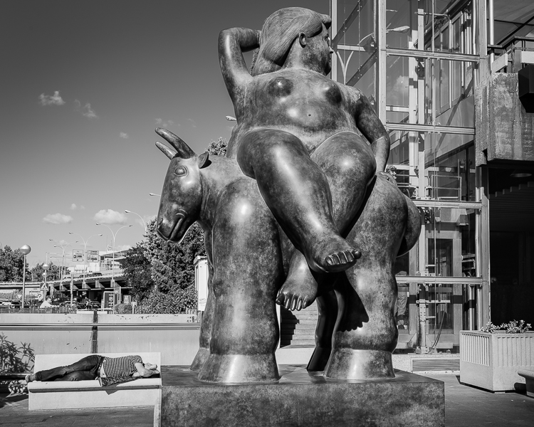Man asleep on bench next to Botero statue outside airport